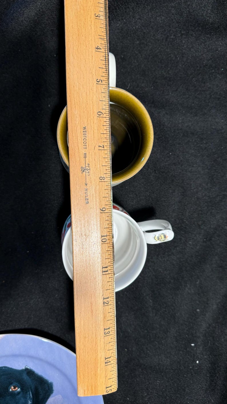 Puede incluir: Dos tazas de cer&aacute;mica con asas blancas y una regla de madera. Una taza es blanca con un dise&ntilde;o rojo y azul, la otra es marr&oacute;n. La regla indica que las tazas miden unos 10 cm de alto.