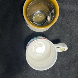 Puede incluir: Dos tazas de cer&aacute;mica, una con interior amarillo y asa blanca, la otra blanca con borde decorativo. Las tazas est&aacute;n sobre un fondo oscuro. Un plato con una ilustraci&oacute;n de perro es parcialmente visible.