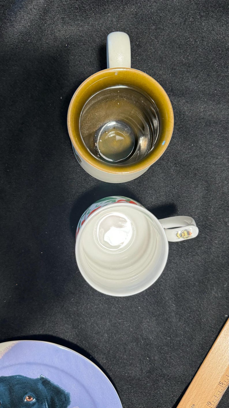 Puede incluir: Dos tazas de cer&aacute;mica, una con interior amarillo y asa blanca, la otra blanca con borde decorativo. Las tazas est&aacute;n sobre un fondo oscuro. Un plato con una ilustraci&oacute;n de perro es parcialmente visible.