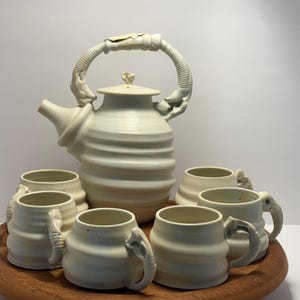 May include: A white ceramic teapot with a unique, sculpted handle and six matching mugs with elephant-shaped handles. The teapot and mugs are arranged on a wooden tray.