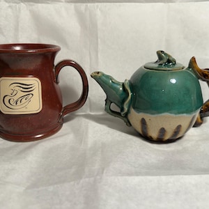 May include: A red ceramic mug with a white logo and a green ceramic teapot with a frog on the lid and a brown handle.