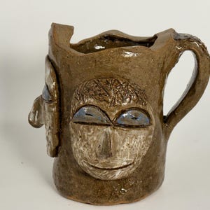 May include: A brown ceramic mug with two faces sculpted on the front. The faces have blue eyes and a textured surface.