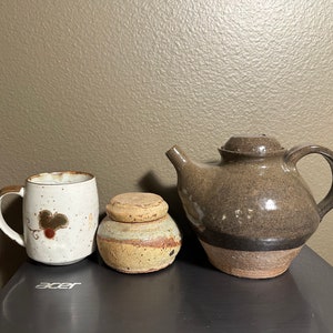 May include: A set of three brown and white ceramic pottery pieces: a teapot, a mug with a brown leaf design, and a small jar with a lid.