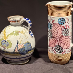 May include: Two ceramic pitchers with hand-painted designs. The smaller pitcher is white with blue, green, and yellow floral patterns. The larger pitcher is white with a pink and brown leaf design and blackberries.