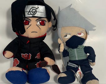 May include: Two plush figures of anime characters. One figure has black hair, a silver headband, and a black outfit with red cloud designs. The other figure has gray hair, a mask, and a green vest over a dark blue outfit. Both figures have detailed facial features.