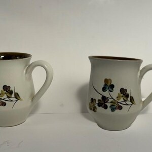 Puede incluir: Dos tazas de cer&aacute;mica blanquecina con borde y asa marrones. Cada taza presenta un dise&ntilde;o floral con hojas verdes, azules y marrones. Las tazas est&aacute;n sobre un fondo blanco liso.