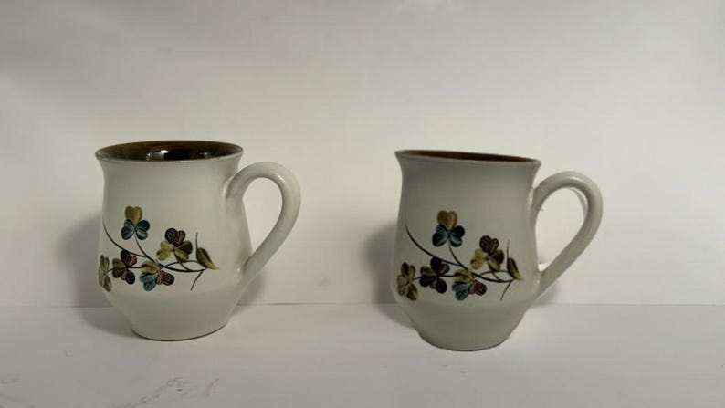 Puede incluir: Dos tazas de cer&aacute;mica blanquecina con borde y asa marrones. Cada taza presenta un dise&ntilde;o floral con hojas verdes, azules y marrones. Las tazas est&aacute;n sobre un fondo blanco liso.