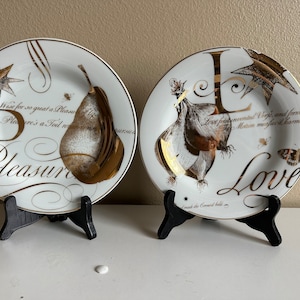 May include: Two white ceramic plates with gold trim and a gold design. The plates feature the letters "P" and "L" with a pear and onion design. The plates have the words "Pleasure" and "Love" written on them.