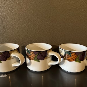 May include: Three white ceramic mugs with a colorful autumnal design featuring brown, orange, green, and purple accents. The mugs have a rounded shape and a handle on the right side.