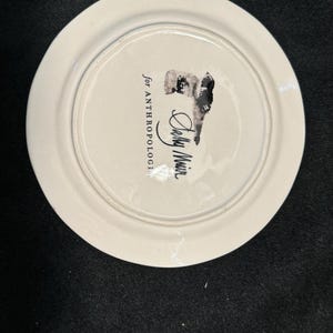 Puede incluir: Plato de cer&aacute;mica blanco con una ilustraci&oacute;n en blanco y negro y el texto "for ANTHROPOLOGIE" y "Sally Muir". El plato tiene un borde ligeramente elevado y un acabado brillante. La ilustraci&oacute;n parece ser un retrato.