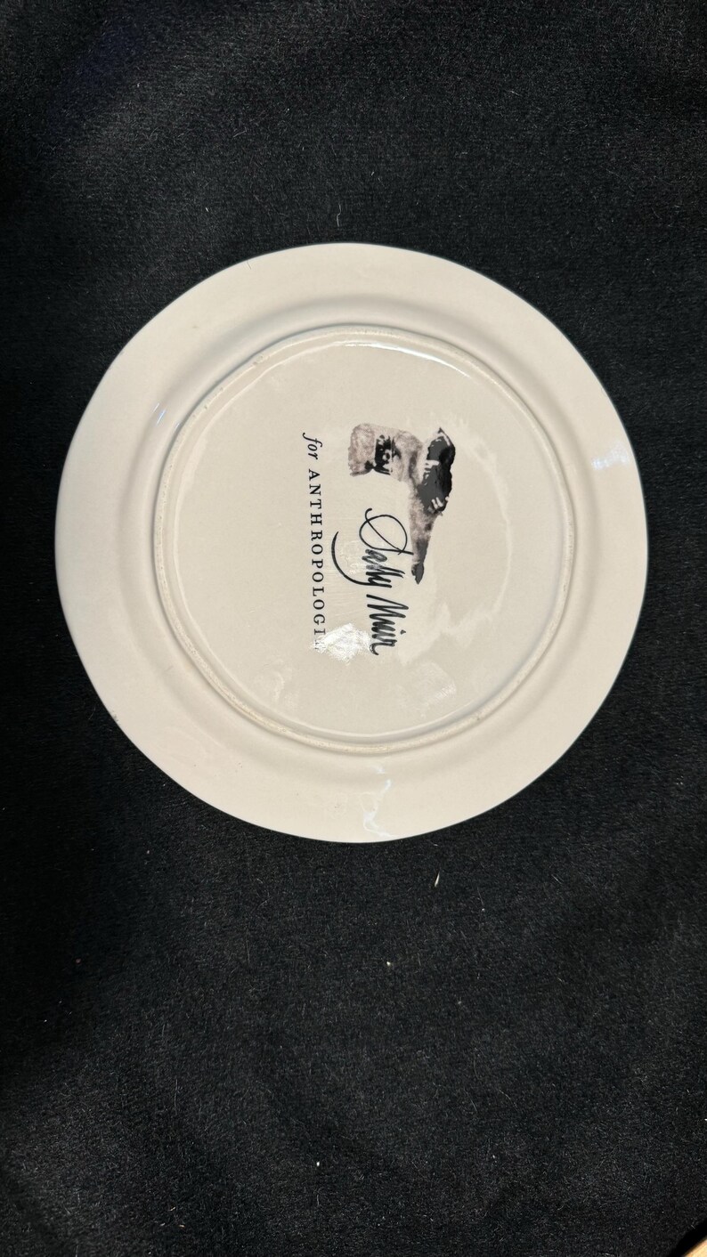 Puede incluir: Plato de cer&aacute;mica blanco con una ilustraci&oacute;n en blanco y negro y el texto "for ANTHROPOLOGIE" y "Sally Muir". El plato tiene un borde ligeramente elevado y un acabado brillante. La ilustraci&oacute;n parece ser un retrato.