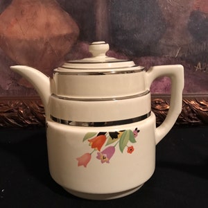 May include: A white ceramic teapot with a floral design featuring orange, pink, and purple flowers. The teapot has a silver trim around the lid and the top of the body.