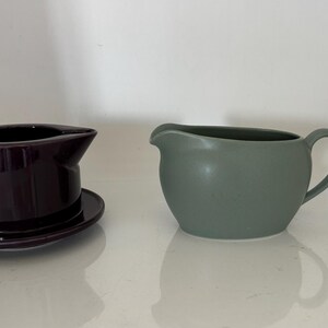 May include: Two ceramic serving pieces. On the left, a dark purple gravy boat with a matching saucer. On the right, a sage green pitcher with a handle. Both pieces are on a white surface.