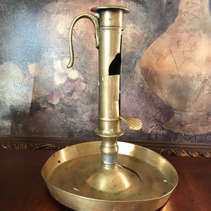 May include: Antique brass candlestick holder with a handle and a circular base. The candlestick has a tall, cylindrical body with a small, curved handle and a small lever. The base is wide and shallow, designed to catch wax drippings.