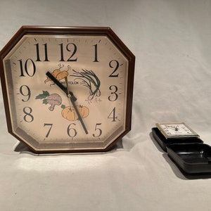 May include: A brown octagonal clock with a white face and black numbers. The clock has a picture of vegetables on the face. The clock is sitting on a white surface. There is a smaller black clock in the lower right corner of the image.