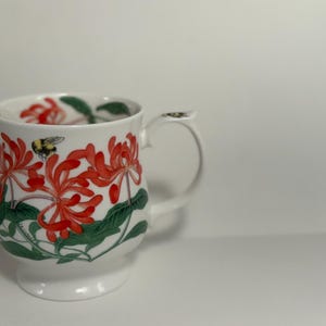Puede incluir: Taza de cer&aacute;mica blanca con un dise&ntilde;o floral de flores rojas, hojas verdes y un abejorro. La taza tiene un asa y una base. El estampado floral envuelve la taza. Un art&iacute;culo decorativo para la cocina o el comedor.