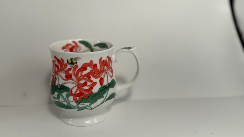 Puede incluir: Taza de cer&aacute;mica blanca con un dise&ntilde;o floral de flores rojas, hojas verdes y un abejorro. La taza tiene un asa y una base. El estampado floral envuelve la taza. Un art&iacute;culo decorativo para la cocina o el comedor.