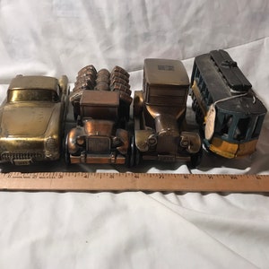 May include: Four vintage metal toy cars, including a green and yellow trolley, a copper truck, a gold car, and a gold classic car. The cars are arranged in a row on a white surface.