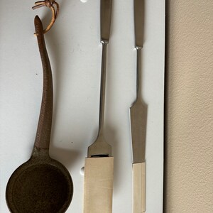 May include: Three metal tools, two with wooden handles and one with a leather strap. The tools are on a white surface.