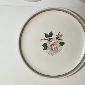 May include: A round, off-white ceramic plate featuring a central floral illustration of a blooming rose with green leaves and a small rosebud. The plate has a smooth, slightly raised rim and a subtle, light-colored border around the edge.