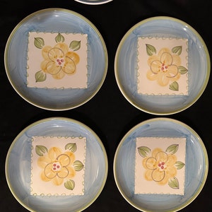 May include: Four blue and white ceramic dinner plates with a yellow flower design in the center. The flowers have a yellow center with red dots and green leaves.