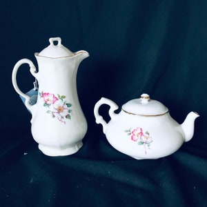May include: A white porcelain teapot and creamer set with floral designs. The teapot has a gold rim and a floral pattern with pink and white flowers. The creamer has a similar floral design.