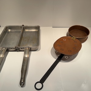 May include: A set of vintage kitchen tools, including a divided metal pan with handles, a copper strainer with a long black handle, and a copper sieve. The tools are arranged on a white surface.