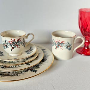 May include: A cream-colored teacup and saucer set with a matching mug, featuring a festive holly and ribbon design. A red glass goblet adds a pop of color to the arrangement. The set includes a dinner plate, a salad plate, and a teacup saucer.