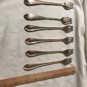 May include: Set of five silver-plated cocktail forks with ornate handles. The forks are laid out in a row on a white fabric surface with a wooden ruler.