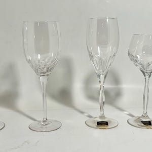 May include: Four clear crystal wine glasses with varying bowl shapes and stem designs. The glasses are arranged in a row, showcasing their elegant forms. The stems have intricate patterns, and the bases are round. The glasses are on a white surface.