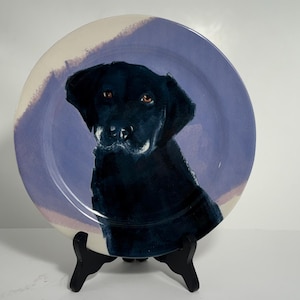 Puede incluir: Plato de cer&aacute;mica decorativo con un retrato de un perro Labrador Retriever negro. El perro est&aacute; pintado con detalle sobre un fondo lila claro y crema. El plato se exhibe sobre un soporte negro.