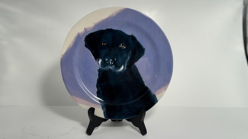 Puede incluir: Plato de cer&aacute;mica decorativo con un retrato de un perro Labrador Retriever negro. El perro est&aacute; pintado con detalle sobre un fondo lila claro y crema. El plato se exhibe sobre un soporte negro.