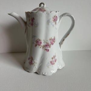 May include: White porcelain coffee pot with a lid and handle. The pot is decorated with pink floral patterns and a scalloped edge. The handle has a distressed, aged appearance.