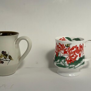 Puede incluir: Dos tazas de cer&aacute;mica. Una es blanquecina con borde marr&oacute;n y dise&ntilde;o floral. La otra es blanca con estampado floral rojo y hojas verdes. Ambas tazas tienen asas.