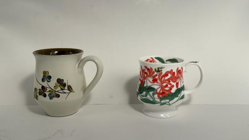 Puede incluir: Dos tazas de cer&aacute;mica. Una es blanquecina con borde marr&oacute;n y dise&ntilde;o floral. La otra es blanca con estampado floral rojo y hojas verdes. Ambas tazas tienen asas.