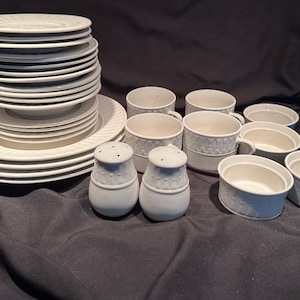 May include: A set of white ceramic dinnerware, including plates, bowls, and cups. The set features a raised, embossed design on the edges of the pieces. There are also two white ceramic salt and pepper shakers.