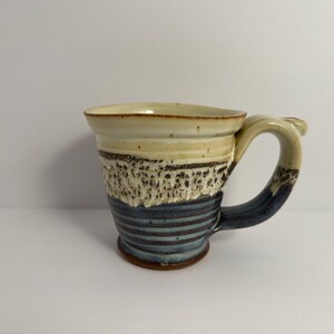 May include: A handmade ceramic mug with a white, brown, and blue glaze. The mug has a wide, rounded base and a handle that curves up and out.
