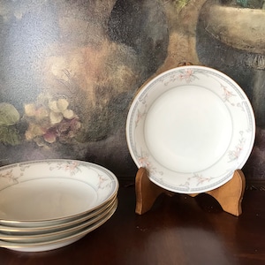 May include: Four white ceramic soup bowls with a floral design in shades of pink and gray. The bowls are stacked on top of each other and are sitting on a wooden table.