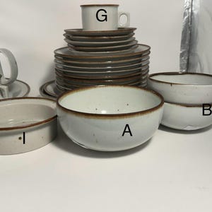 May include: A collection of off-white ceramic tableware with brown trim. The set includes bowls, plates, cups, and a serving dish. The pieces are stacked and arranged on a white surface, showcasing a complete dining set.
