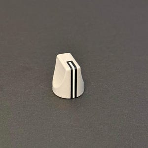 以下が含まれることがあります： 平らな上面と丸みを帯びた側面を持つ、明るい灰色の三角形のノブ。2本の垂直な黒いストライプが前面を走り、その間に白いストライプがあります。ノブは、電子機器用である可能性があります。