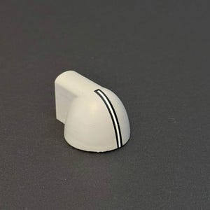May include: A light gray, angled knob with a curved front. A vertical black stripe with a white border accents the curved surface. The knob has a rectangular base and is likely used for appliance control.