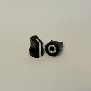 Op de afbeelding: Twee zwarte bedieningsknoppen met een witte indicatorlijn. Eén knop staat rechtop en toont de lijn, terwijl de andere op zijn kant ligt en een zilverkleurig midden onthult. Deze knoppen zijn waarschijnlijk voor elektronische apparaten.