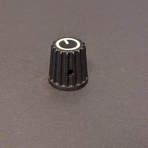 May include: A black control knob with a white indicator line. The knob has a ridged, conical shape and a small hole. The knob is likely used for adjusting settings on electronic equipment.