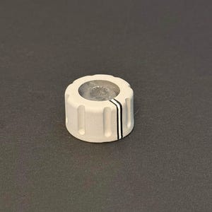 May include: A white control knob with a clear center and vertical grooves. Two black stripes are on the side. The knob is likely used for adjusting settings on electronic devices.