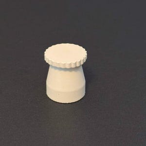May include: A small, off-white, decorative bottle stopper. The stopper has a cylindrical body with a flared base and a scalloped, circular top. The object is made of a solid, opaque material and sits on a dark gray surface.