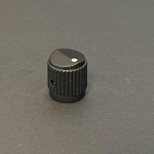 May include: A black control knob with a ridged cylindrical base and a slanted top. The top is divided into two sections, one black and one lighter, with a small white dot. The knob is likely used for adjusting settings on electronic devices.