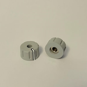 Op de afbeelding: Twee lichtgrijze bedieningsknoppen met een cilindrische vorm. Elke knop heeft een centraal gat en een witte streep met zwarte randen. De knoppen zijn ontworpen voor elektronische apparaten en hebben een metalen inzetstuk.