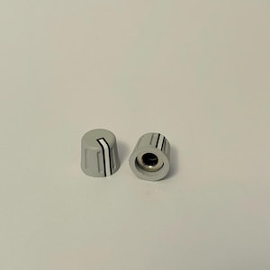 Op de afbeelding: Twee grijze bedieningsknoppen met een witte streep en twee zwarte strepen. De knoppen zijn cilindrisch met een licht taps toelopende bovenkant. Eén staat rechtop, de andere ligt op zijn kant en toont een centraal gat.