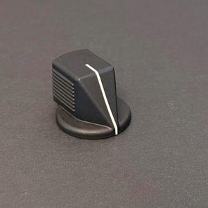May include: A black control knob with a white stripe and a ridged side. The knob has a circular base and a slanted top. The knob is likely used for adjusting settings on electronic devices.