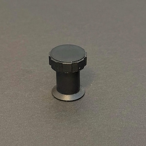 May include: A black, cylindrical object with a flat, circular top and a ridged edge. The object has a short, straight stem and a wider, flat base. The item is set against a dark gray background.
